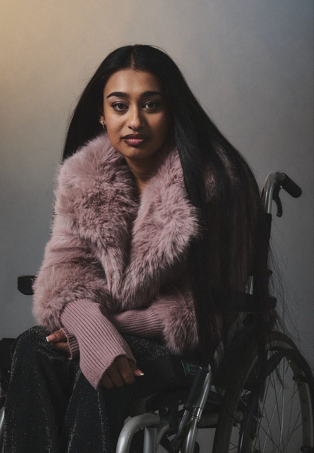 A southeast Asian person with long black hair, wearing a dusty-pink faux-fur jacket and using a wheelchair, pictured against a grey gradient background.