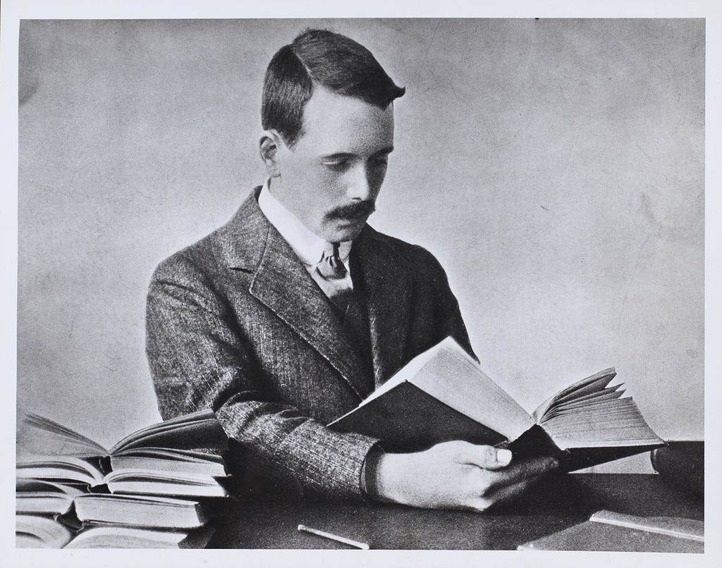 Black and white photograph of Henry Gwyn Jeffreys Moseley seated reading a book, with many open books at his side.