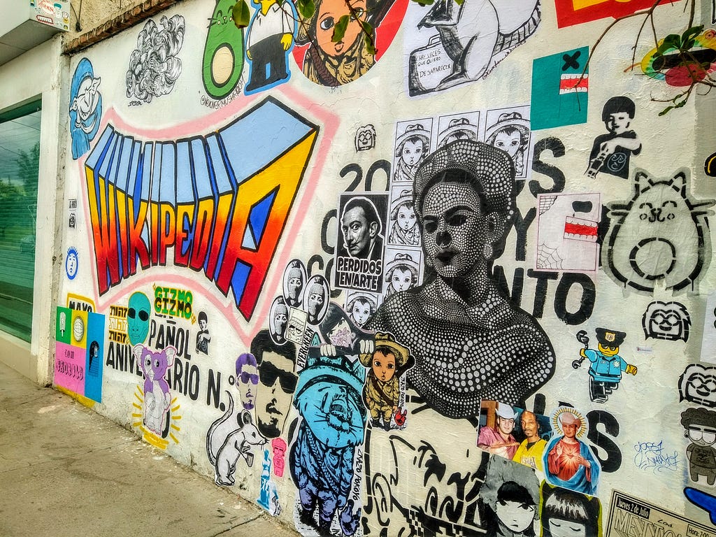 A photograph of a Wikipedia mural intervened by stickers, posters, and stencils in Aguascalientes, Mexico