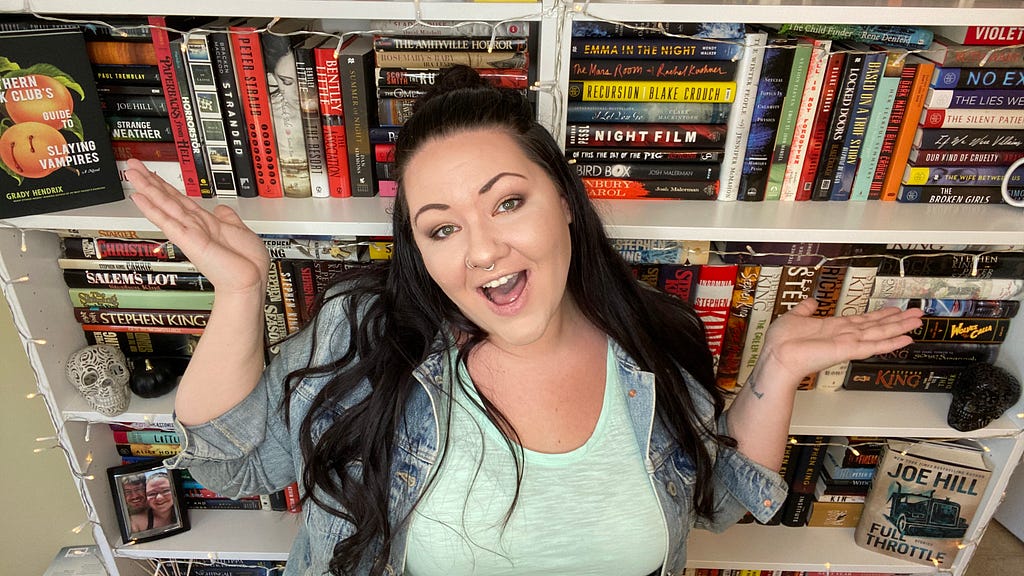 Myself with my hands up, smiling, standing in front of my white bookshelves, full of books.