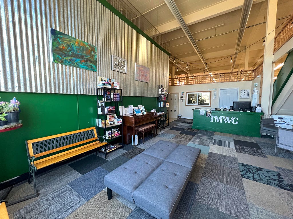 The interior of The Moore Wright Group’s Recovery Café of Hope. The lower walls and welcome counter are dark green with a collection of benches and bookshelves throughout. Paintings by TMWG staff hang on the walls.