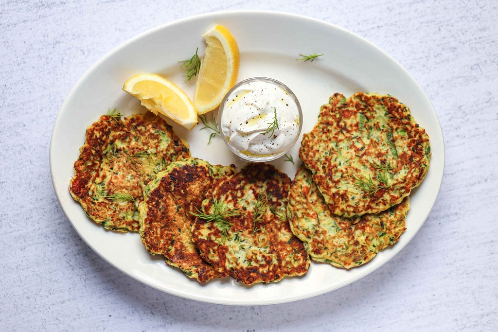 Plate of zucchini pancakes and Greek yogurt sauce, garnished with two lemons, by FIT & NU.
