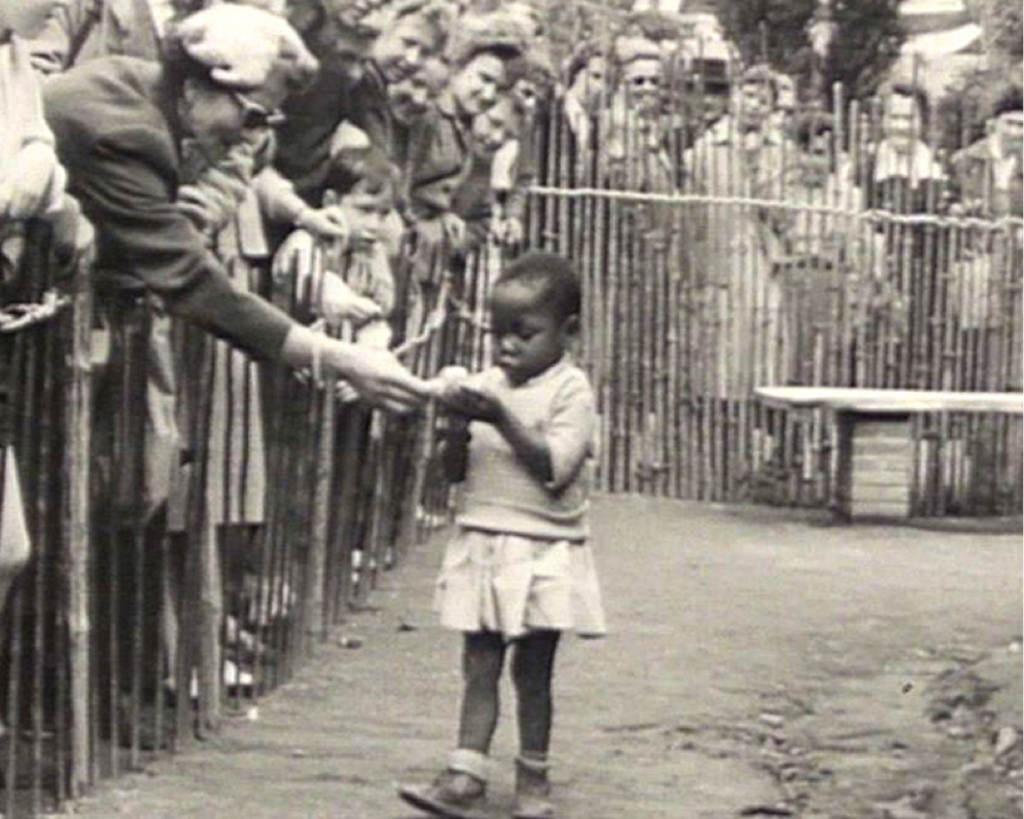 Circa 1904. “Human zoo.” White patrons look from behind a guardrail at a child of African descent. Someone hands them food.
