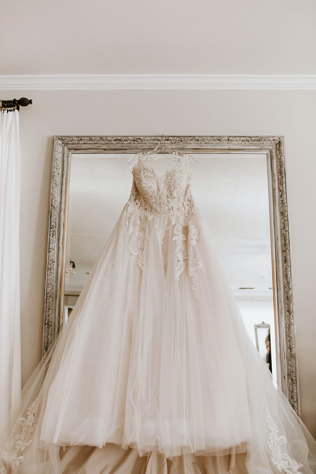 Wedding gown with intricate bodice and flowy, sheer skirt hanging on a large framed mirror against a wall.