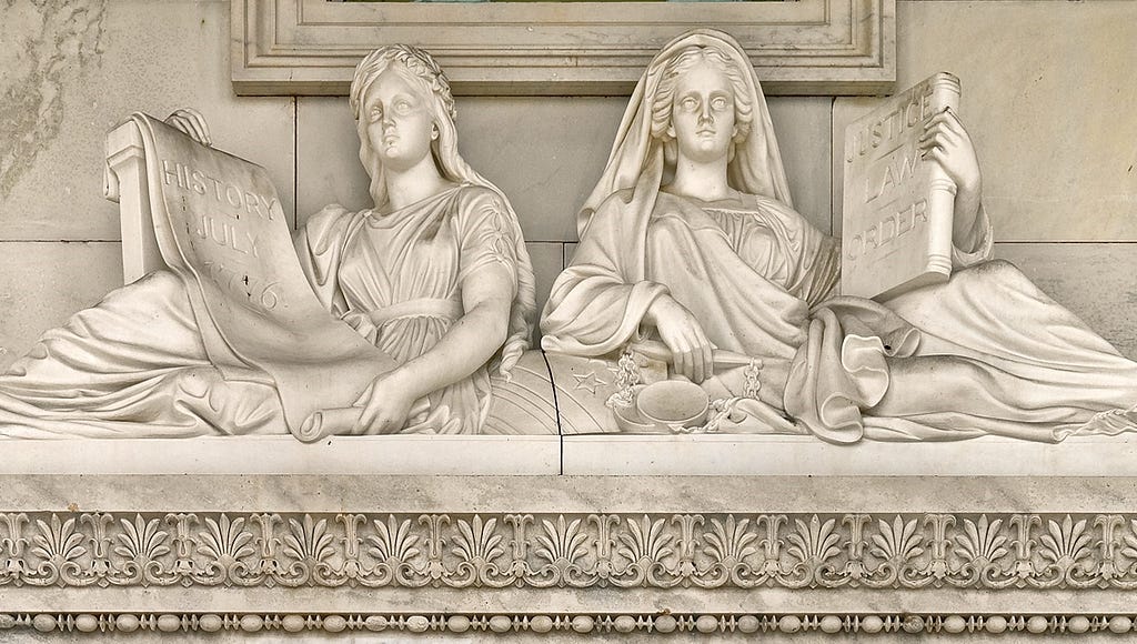 Justice and History marble sculpture in U.S. Capitol features two draped female figures reclining against a globe. Justice holds a book inscribed “Justice / Law / Order” in her left hand; her right hand rests on a pair of scales. History holds a scroll inscribed “History / July / 1776.”