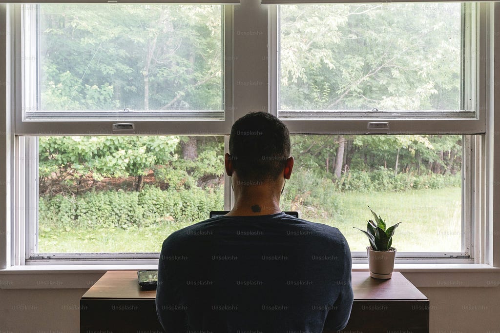 A man looks out of a double window at a wooded area while sitting at a wood desk with a plant on it.