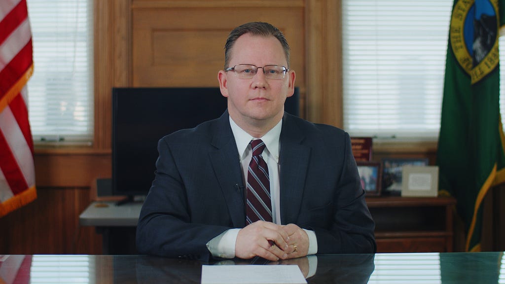 Superintendent Chris Reykdal sitting at his desk with his hands crossed in front of him.