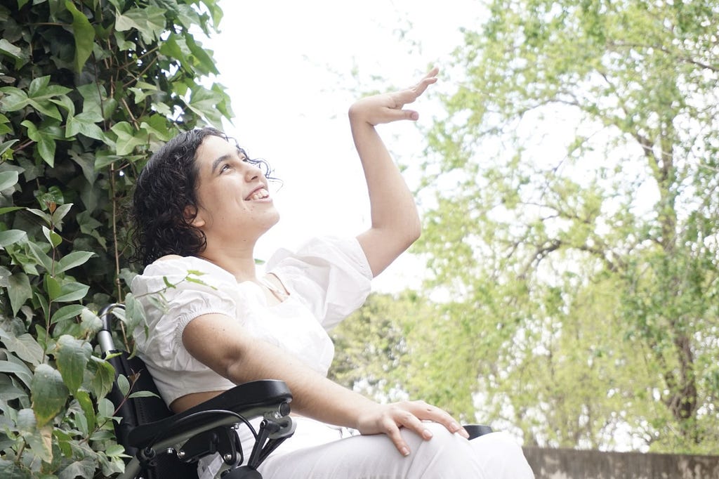 Gianna en su silla de ruedas, vestida de blanco levantando la mano izquierda con un fondo de árboles verdes y cielo nublado