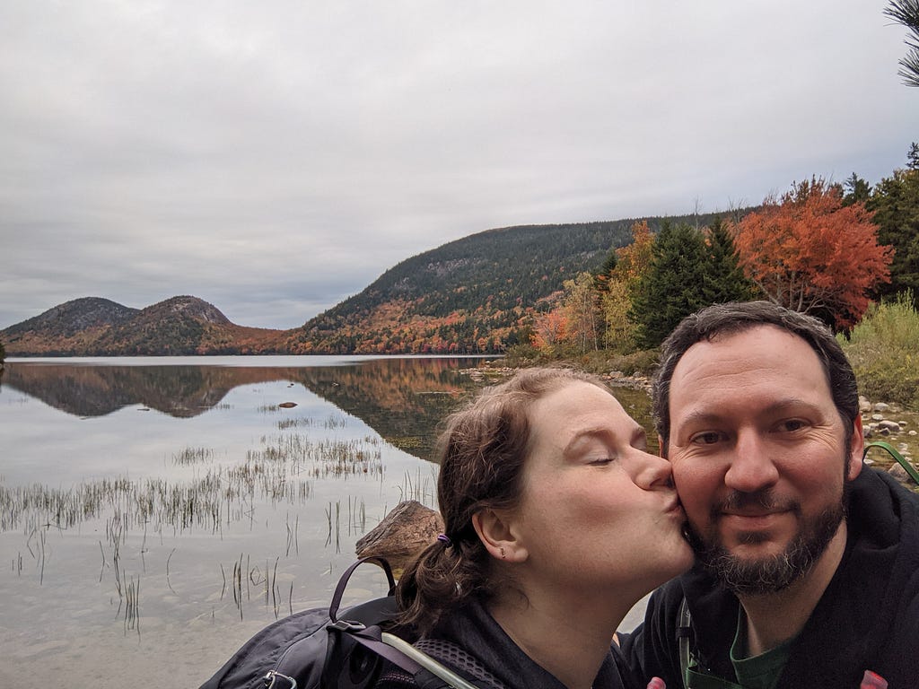 Me kissing hubby’s cheek in front of an overcast vista of a pool reflecting hills in Acadia National Park