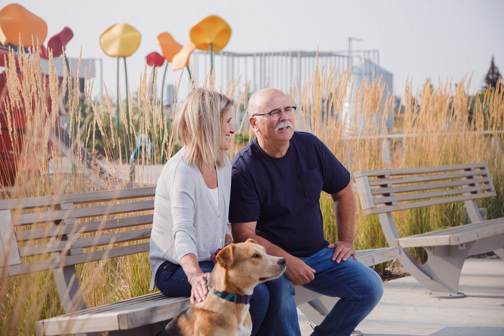 A senior couple pause on a bench with their dog. Behind them, long grasses and a playground sculpture of large metal flowers colour the scenery.