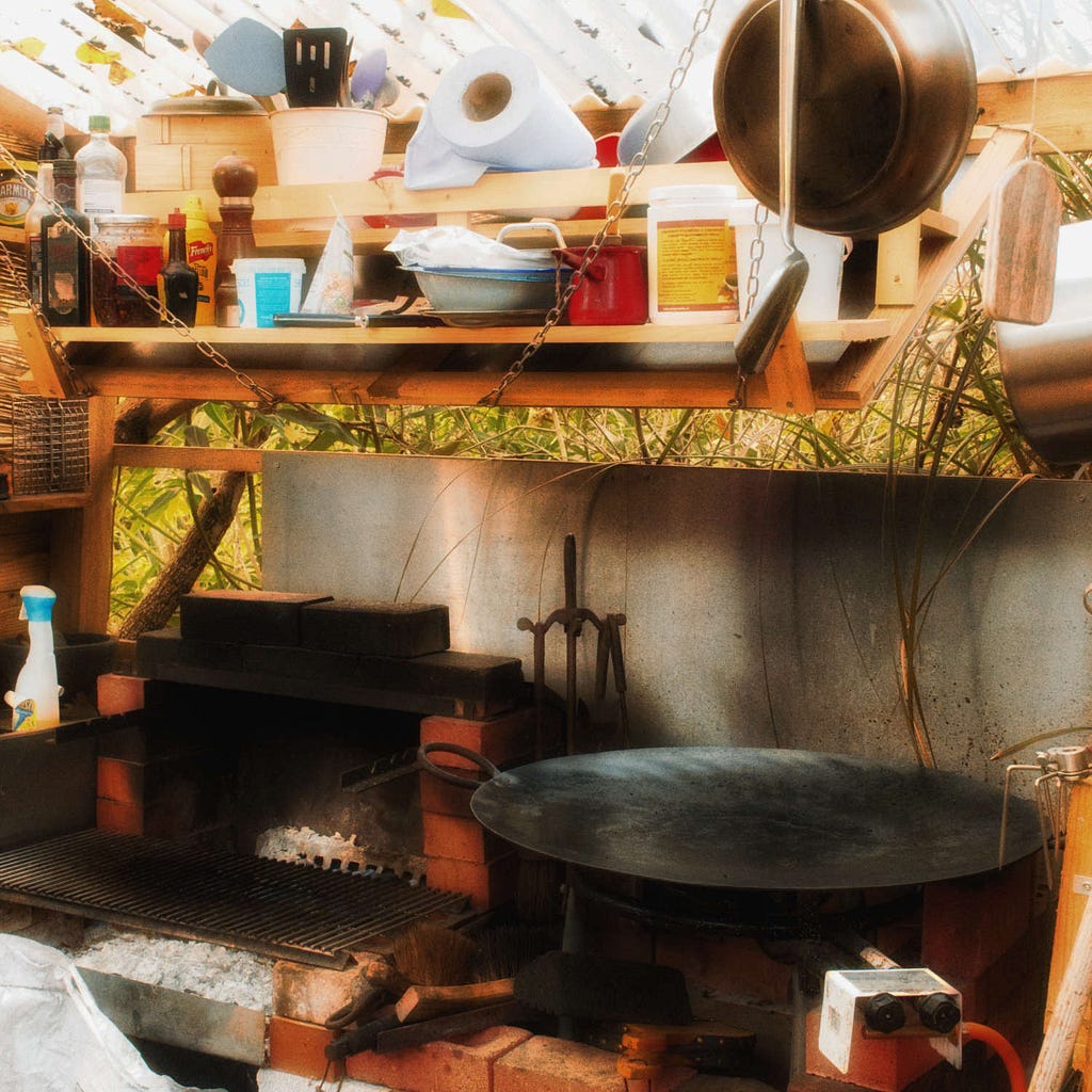 An outdoor kitchen with a charcoal grill on the left, a tawa on the right, and a hood above covered with pans and ingredients