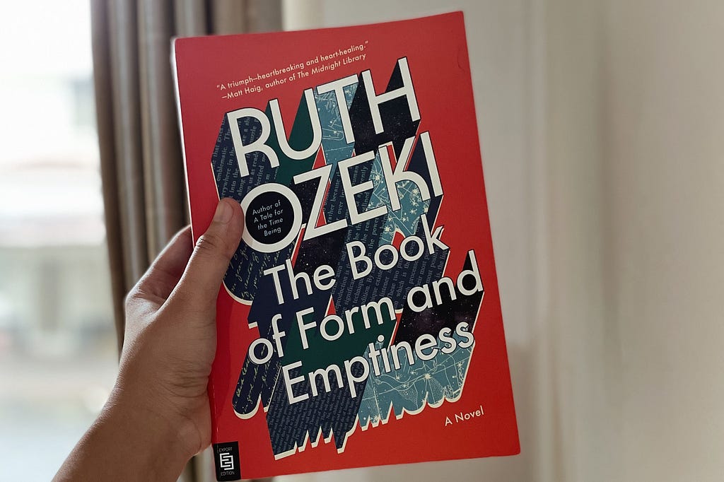 A hand holding a red covered book entitled The Book of Form and Emptiness by Ruth Ozeki