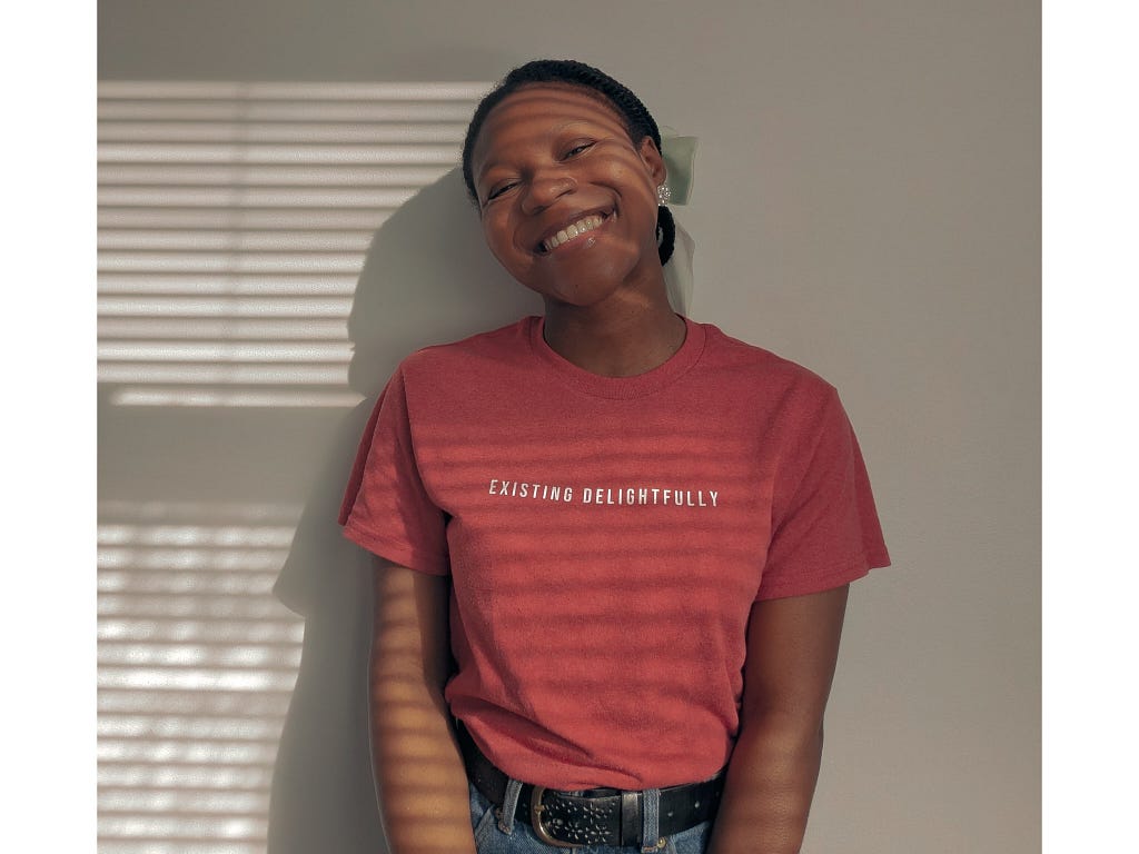A picture of the writer Nnebuugo wearing a T-shirt that says “Existing delightfully” and smiling.