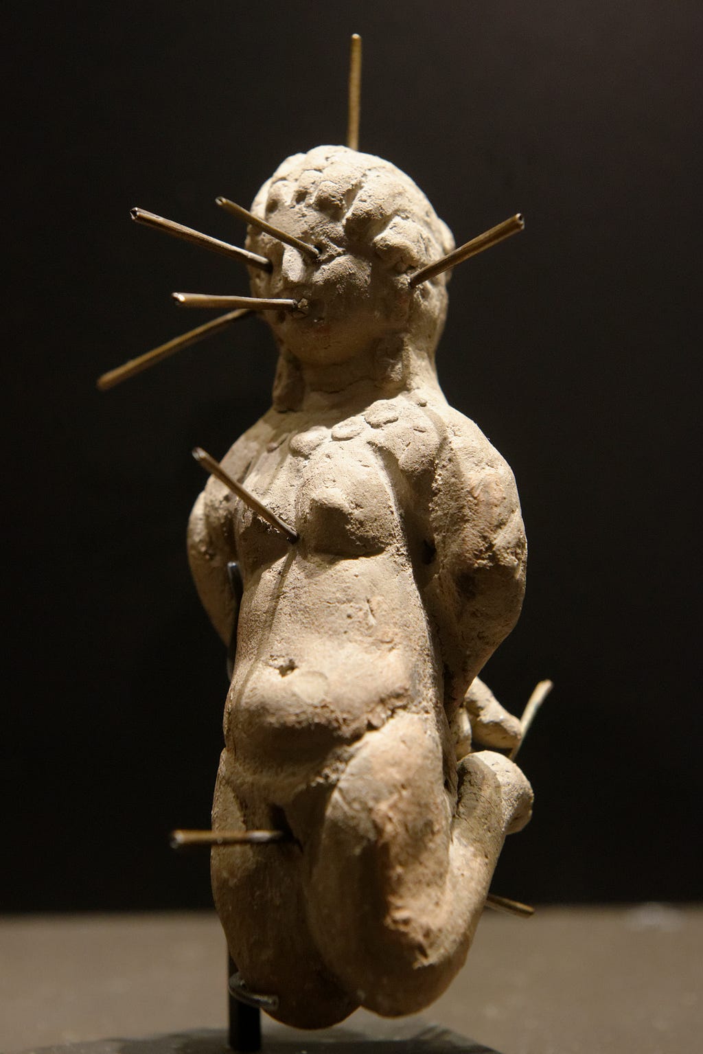 A clay figure is depicted, bound, pierced with needles.