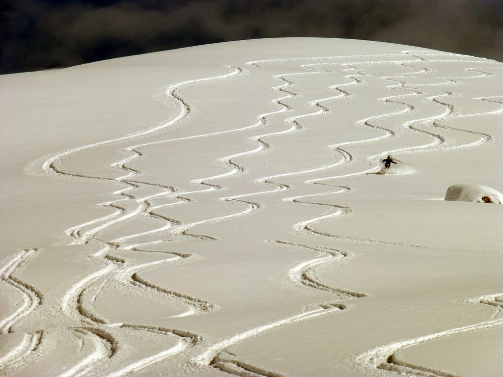 Image shows a skier making their way down an off piste snow track, with snow marks left from other skiers before them
