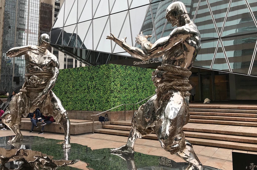 Pictured: Statues of two men in traditional martial arts poses facing one another, ready to fight. Photo taken in front of the Standard Chartered building, Forum, Hong Kong, Central.