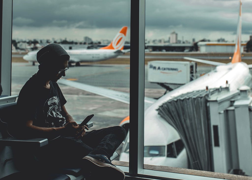 A photo of a person sitting at an airport gate. Taxiing airplanes, jetways, and pavement can be seen through the window behind them.