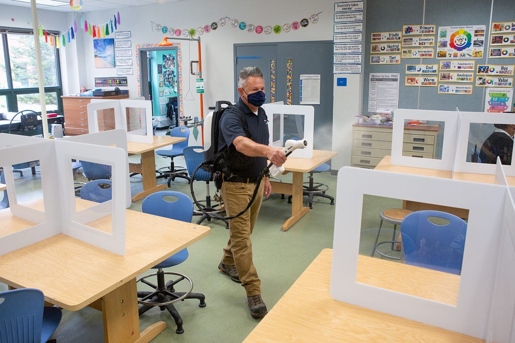 An adult man wearing a face mask sprays disinfectant in an empty classroom.