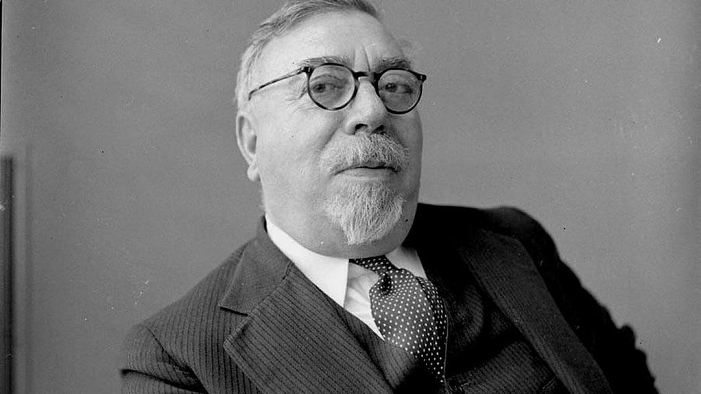 Black-and-white portrait of Norbert Wiener wearing round glasses, a striped suit, vest, and polka-dotted tie. He looks slightly upward with a composed expression, his short beard neatly trimmed.