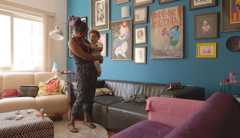 Uma mulher negra carrega um bebê nos braços. Ela está em meio a uma sala com sofá e quadros na parede.