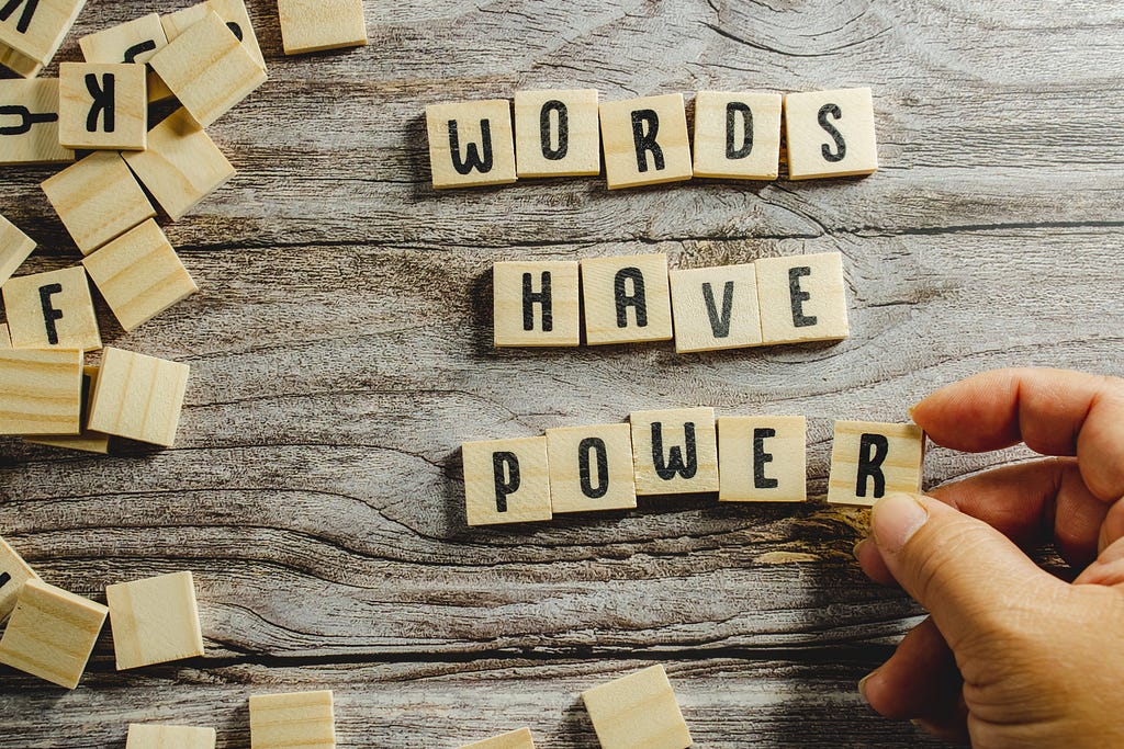 On the left side of the photo, small wooden letter tiles are scattered on a wood grained surface. At center right, tiles form the words “WORDS HAVE POWER,” one word per row. A person’s hand is shown at bottom right placing the last tile in the word “POWER.”