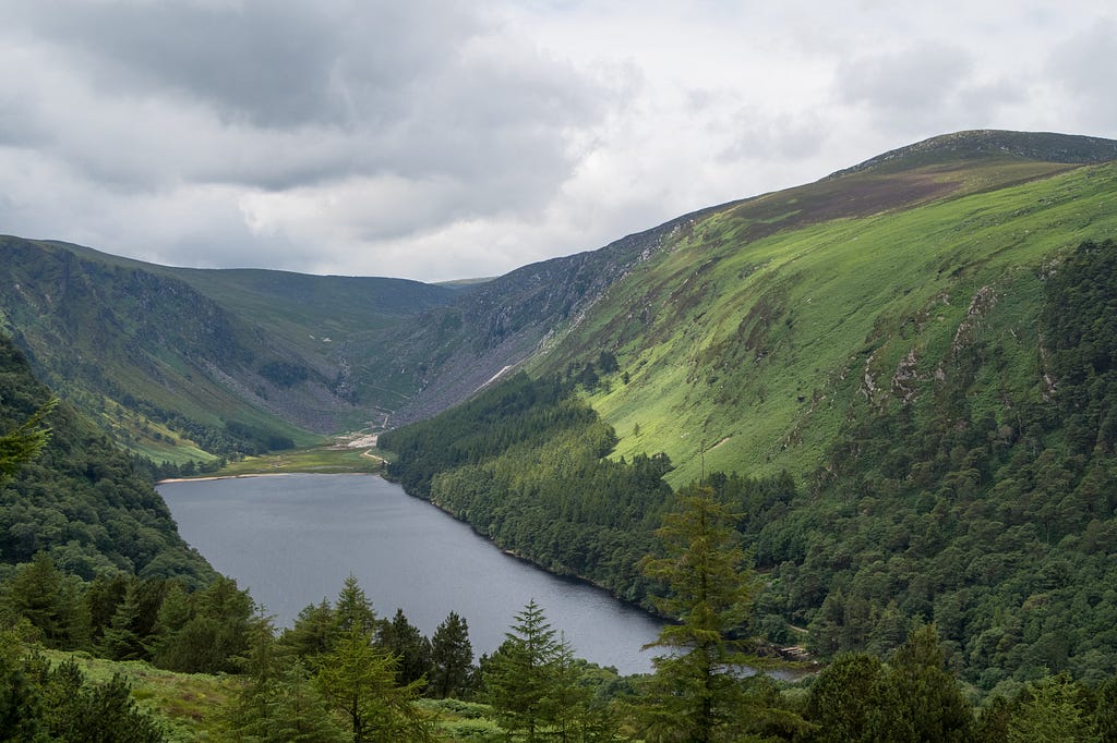 Glendalough with its undulating greenery surrounding a valley lake with a cloudy sky.