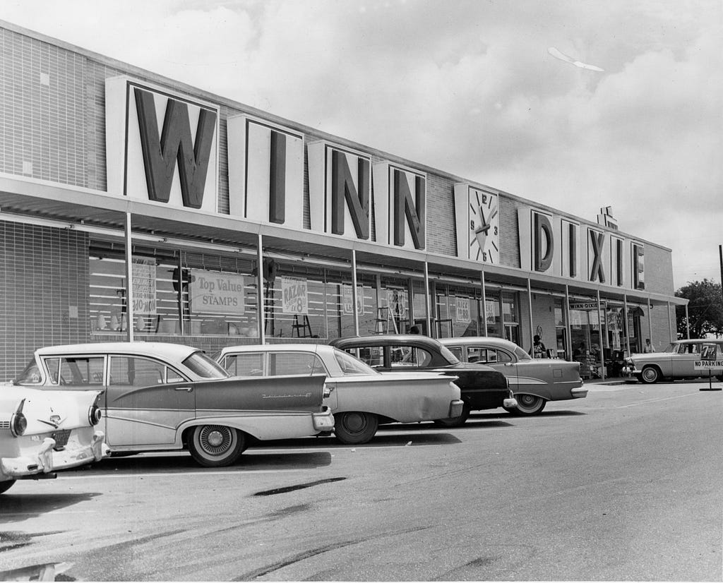 A black and white photo of a Winn-Dixie supermarket and its parking lot which has cars from the 50s parked in front.