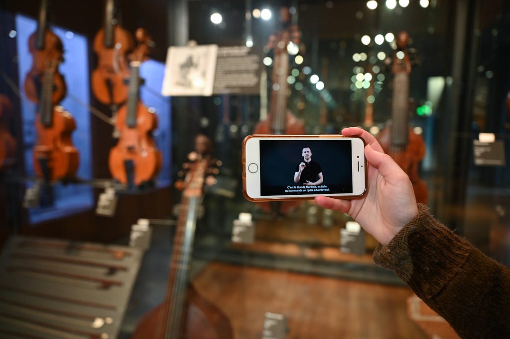 A person’s hand holding a smartphone with a video of a sign language interpreter on its screen. In the blurred background, a display of violins and other string instruments in a museum exhibit, indicating an accessibility feature for deaf visitors.
