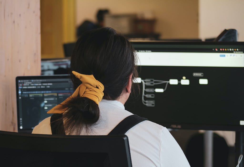 Woman with white long sleeve shirt with back turned and facing a monitor screen