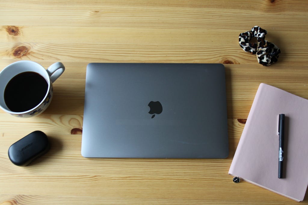 A laptop, a cup of coffee and a notebook sits on a wooden desk.