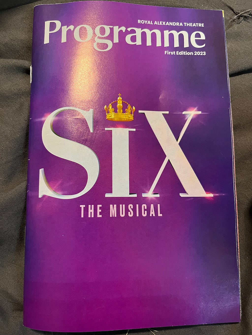 A programme for SIX The Musical. The show’s title is printed in white letters on a purple background, with a gold crown above the I. Additional text in white states that it is a programme, that the show is presented at the Royal Alexandra Theatre, and that it is First Edition 2023.