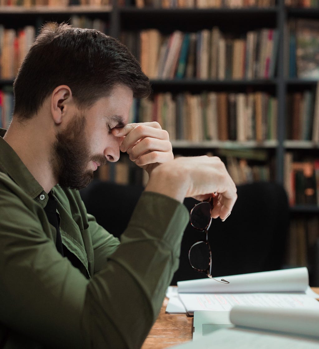 A caucasian man in profile view pinches his nose and holds out his glasses with his free hand. His head is pointing down and his eyes are closed. He is wearing a green collared shirt. He has short brown hair, a beard and a moustache. Below him is a thick wad of paper. Behind him are shelves of books.