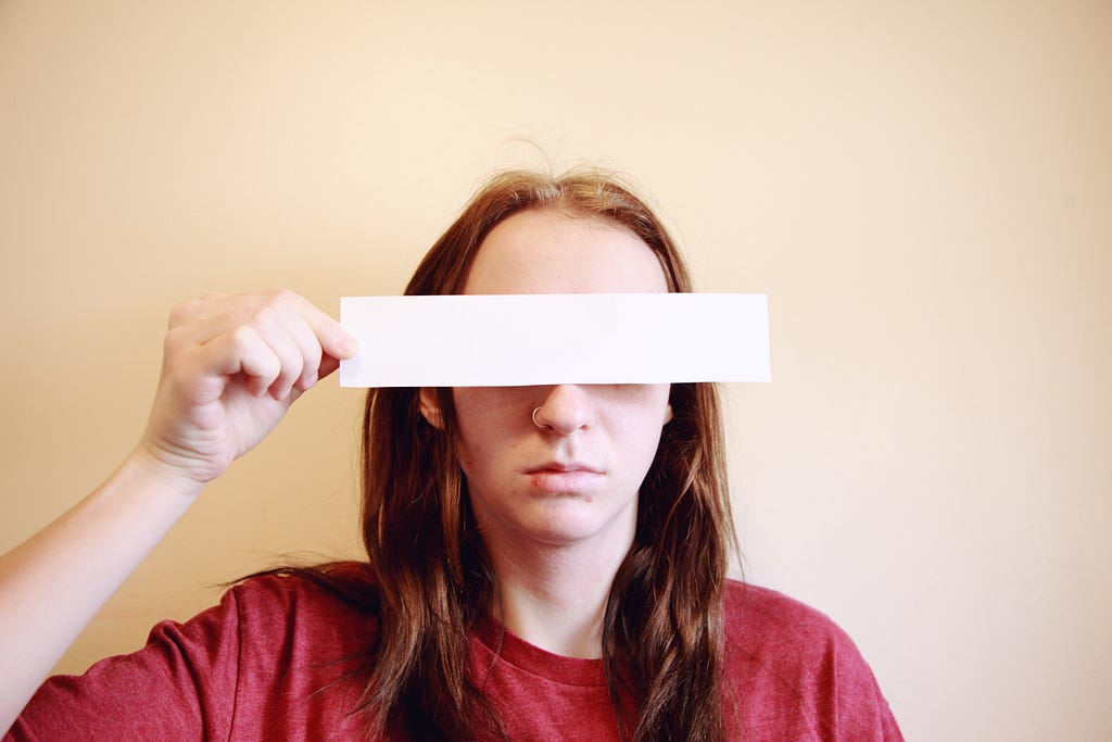 Woman with straight red hair and red shirt holding a rectangular blank piece of paper over her eyes.
