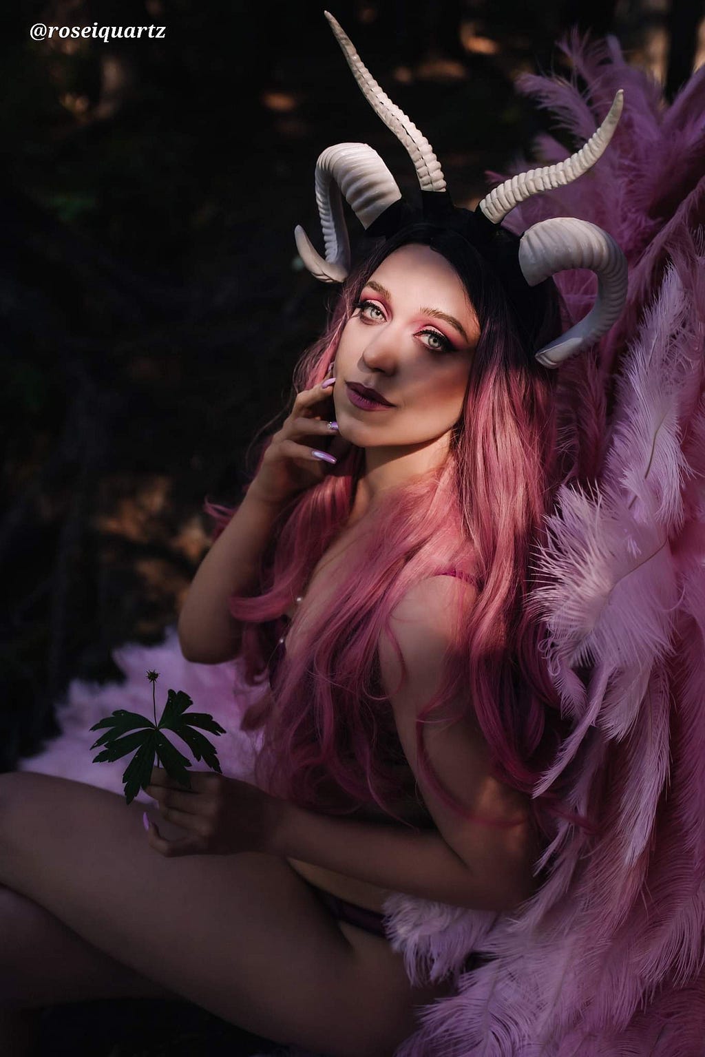 A thin white woman wearing full cosplay, including face make-up, feathered wings, and horns, but not much else, sits in a forest holding a leaf.