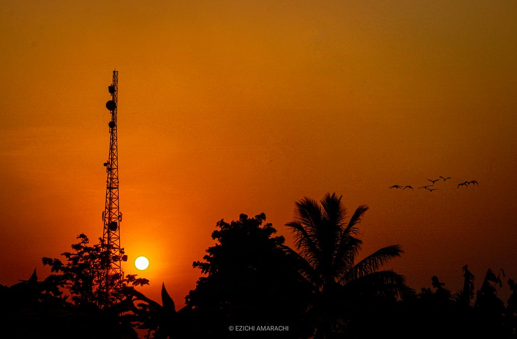 An orange-themed photo of the sun setting. To the left, the sun is very vivid and round and it’s behind a tall communication mast, and to the right, here’s a group of birds flying by. Some trees form a silhouette on the photo. It’s a really beautiful sight.