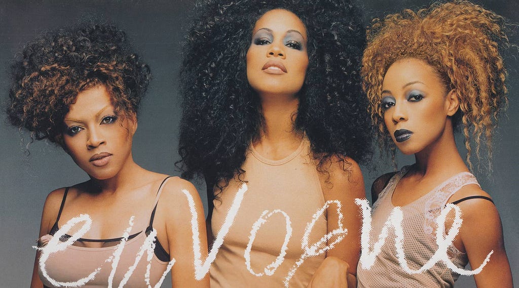 Maxine Jones, Cindy Herron-Bragg, and Terry Ellis sport their outlandish, voluminous, black-and-brown hairstyles from the “Whatever” music video while wearing flesh-toned undergarments. Where the music video stylizes them under sickly green light, this image depicts them as fashionistas in more flattering warm incandescent tones. The image was used as the cover of their follow-up single “Too Gone, Too Long.”