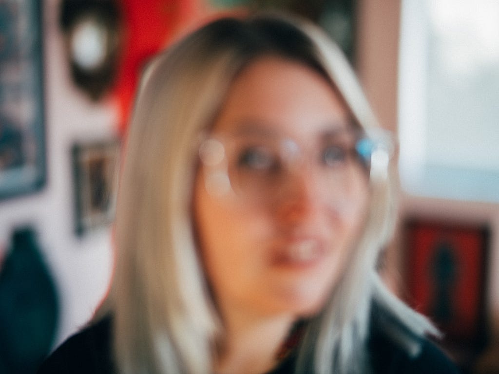 Erin looks ahead, camera focused blurred and fuzzy, slightly off-camera, framed by warm studio tones and subtle pops of red in the background.