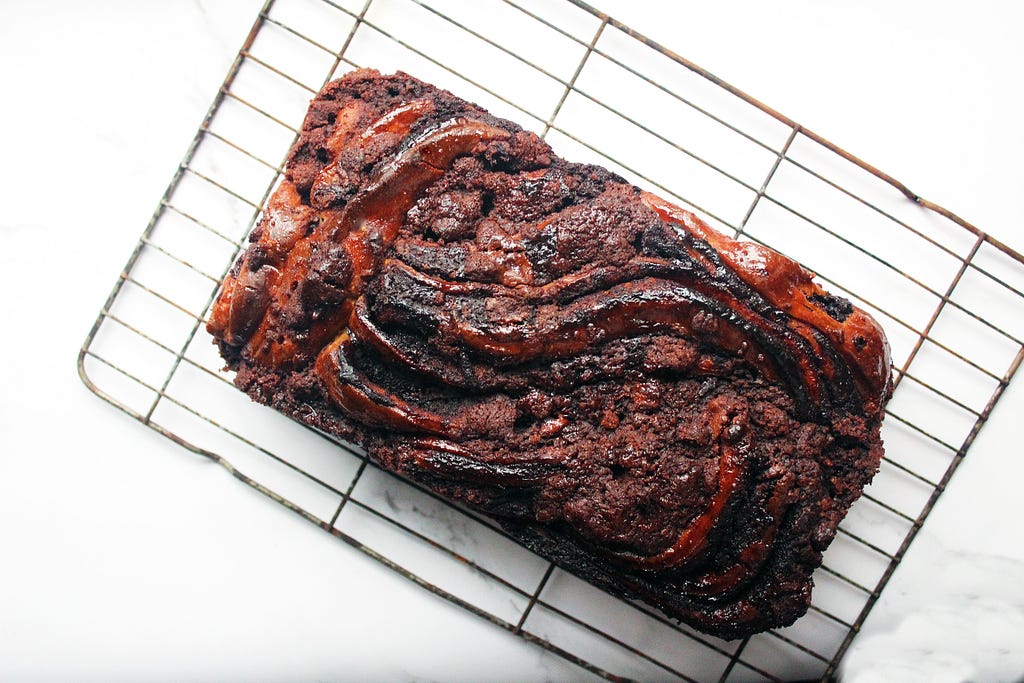 Chocolate babka loaf cake as seen from above. It is resting on a wire rack on a white marble surface.