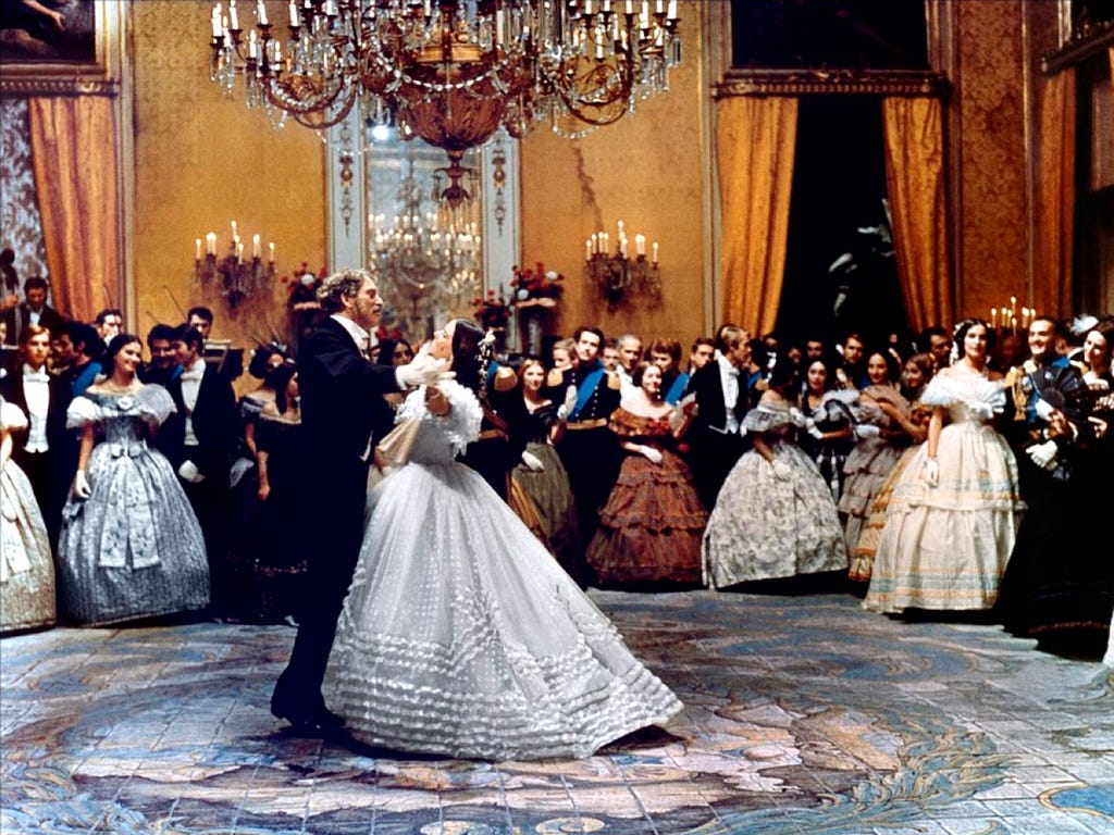 An elegant, mature man and a young woman in a fabulous white gown prepare to lead a dance at a nineteenth-century ball in a luxurious Italian palace
