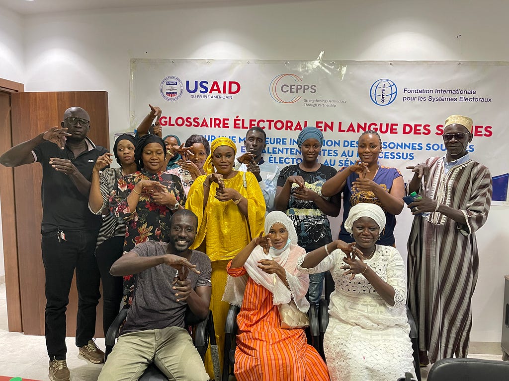 A group of people in front of a USAID banner pose for a picture while using sign language to display the word “vote” with their hands.
