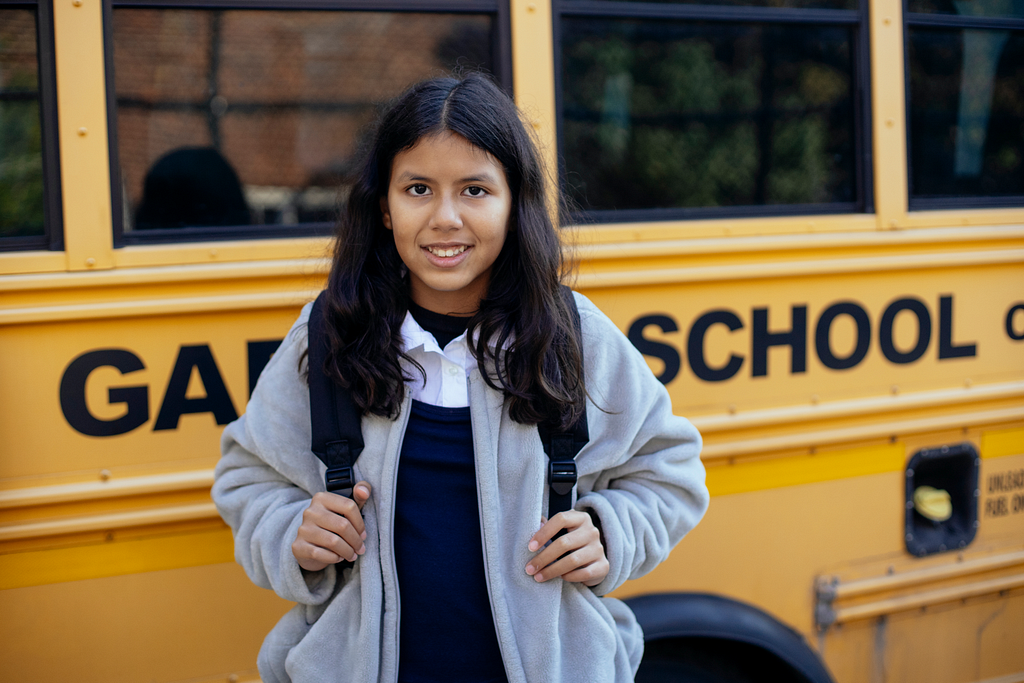 A preteen girl stands smiling in front of a school bus.