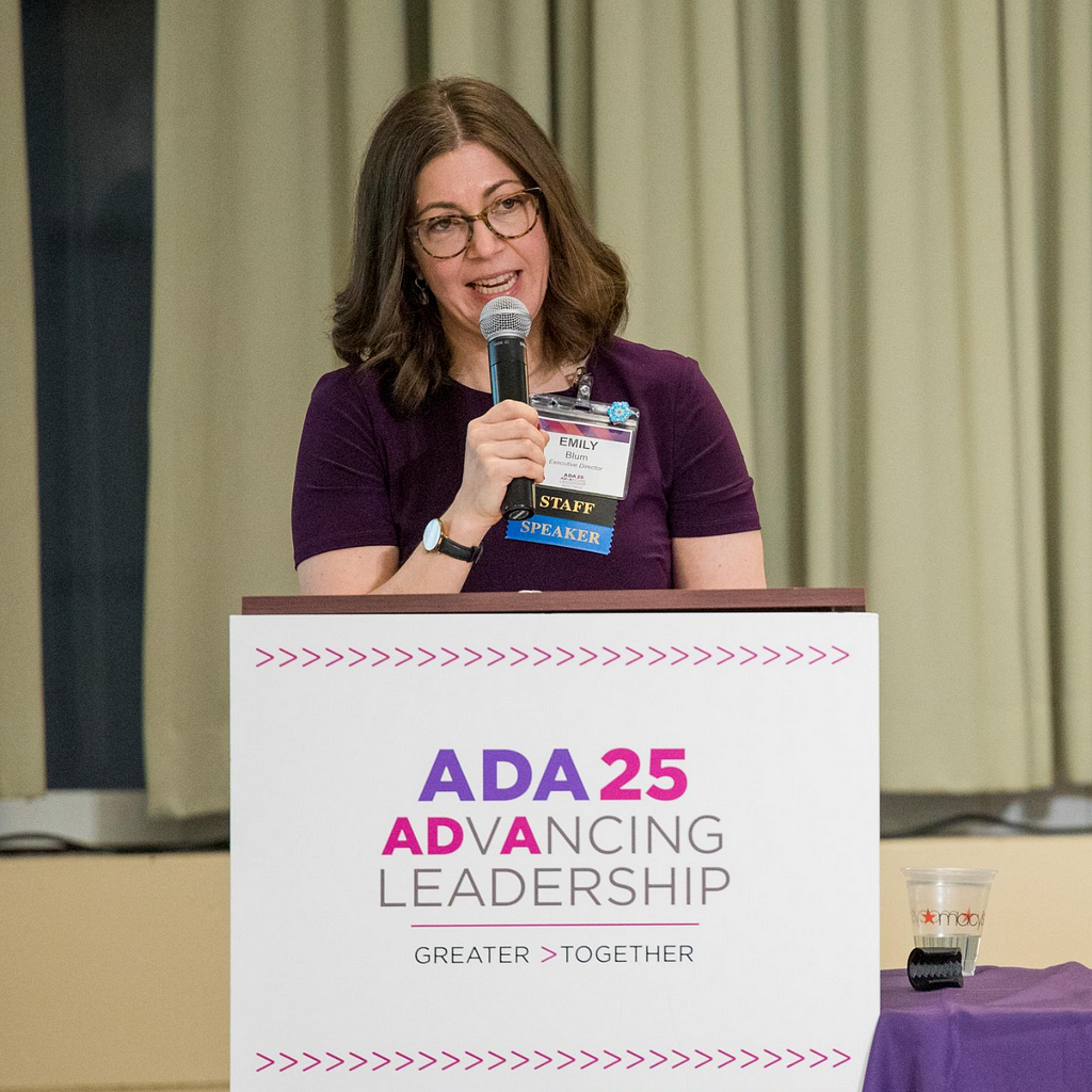 Emily speaks into a mic at a podum with the ADA 25 logo. Brown, medium length hair. Short-sleeved maroon dress, glasses.