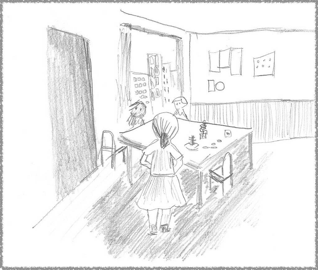 Classroom with a large table at the center and a teacher standing at the edge of the table, facing back from the perspective of the viewer. There are two students on the opposing side of the table and various toys sitting on the table.