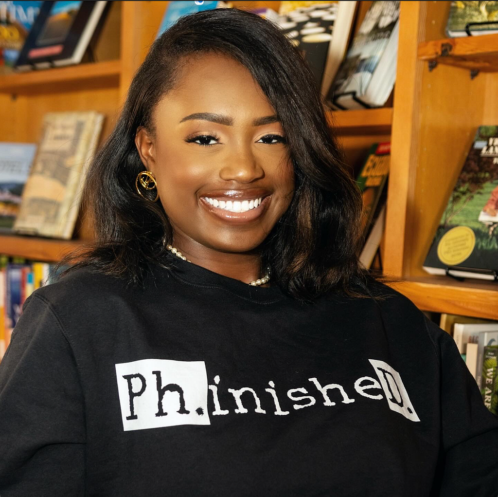 Portrait of Black woman in a library. She is smiling ear-to-ear and wearing a Black crewneck that says Ph.inisheD. in white and black letters. She has wavy black hair with brown highlights.