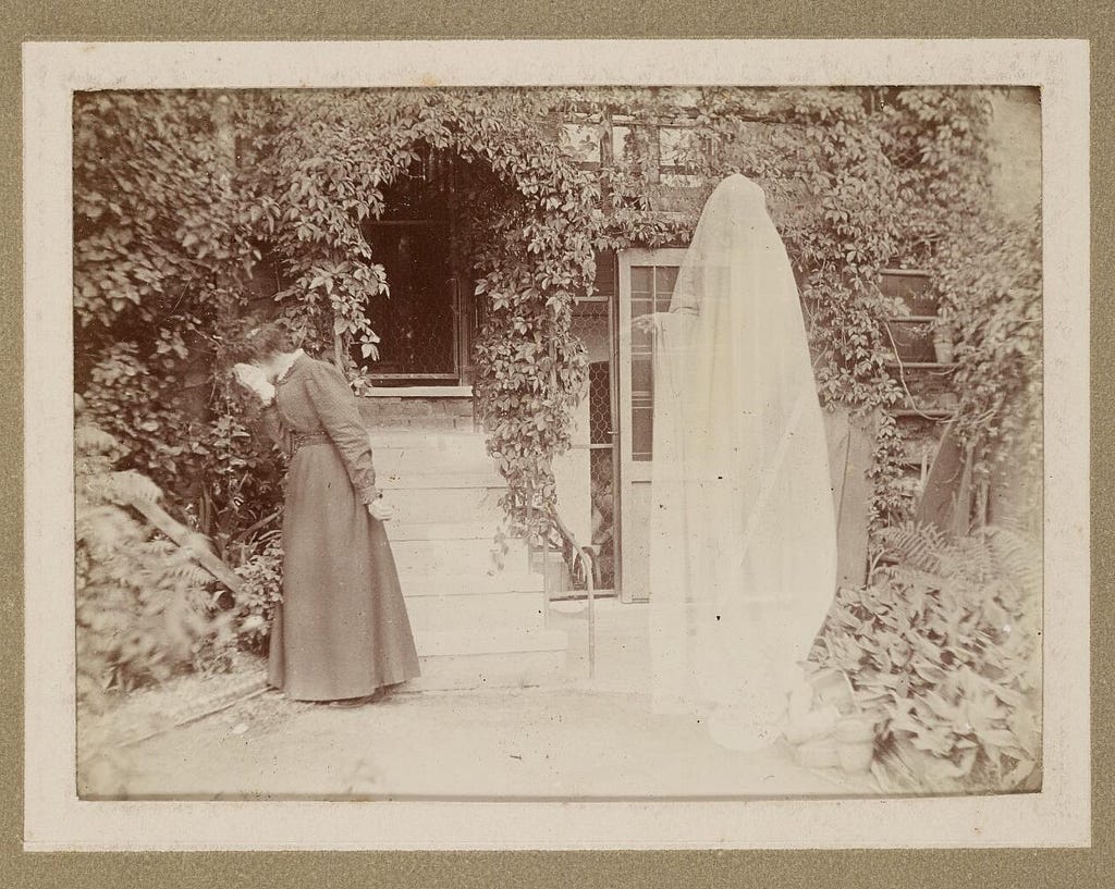 In a lush courtyard, close to a glazed and overgrown building entrance, a woman covers her face and turns away from a white-translucent sheet-covered ghost pointing at her with its singularly visible hand.