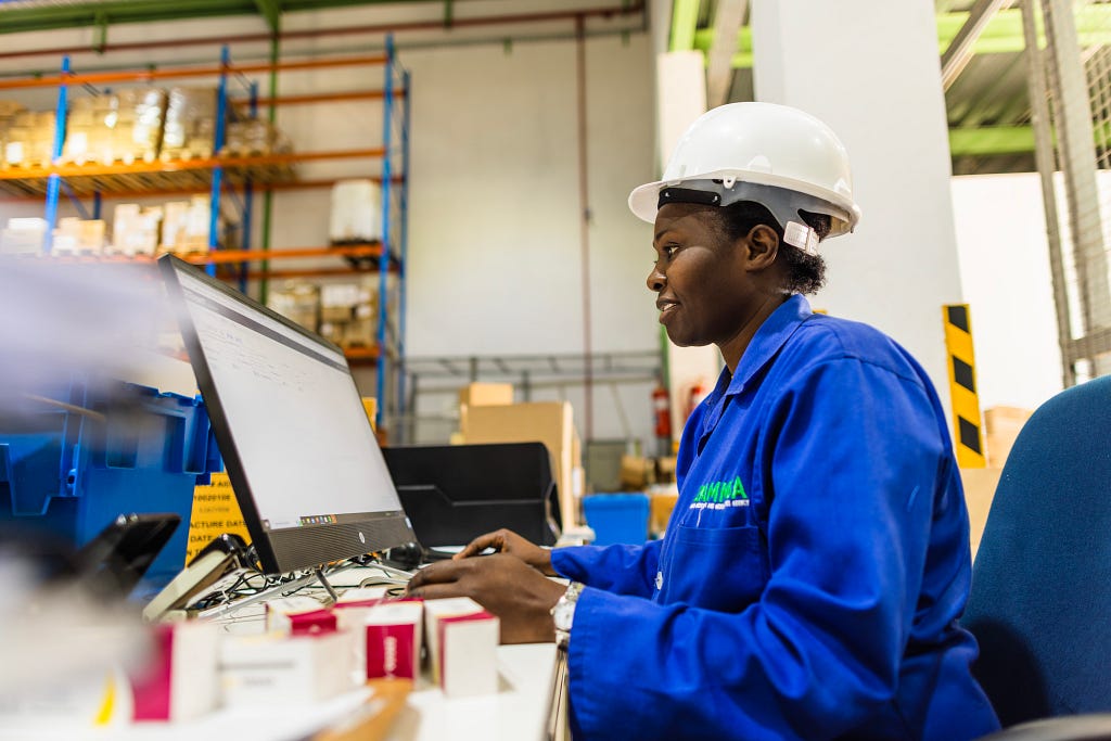 A woman in a white hardhat and blue lab coat inputs information into a computer.