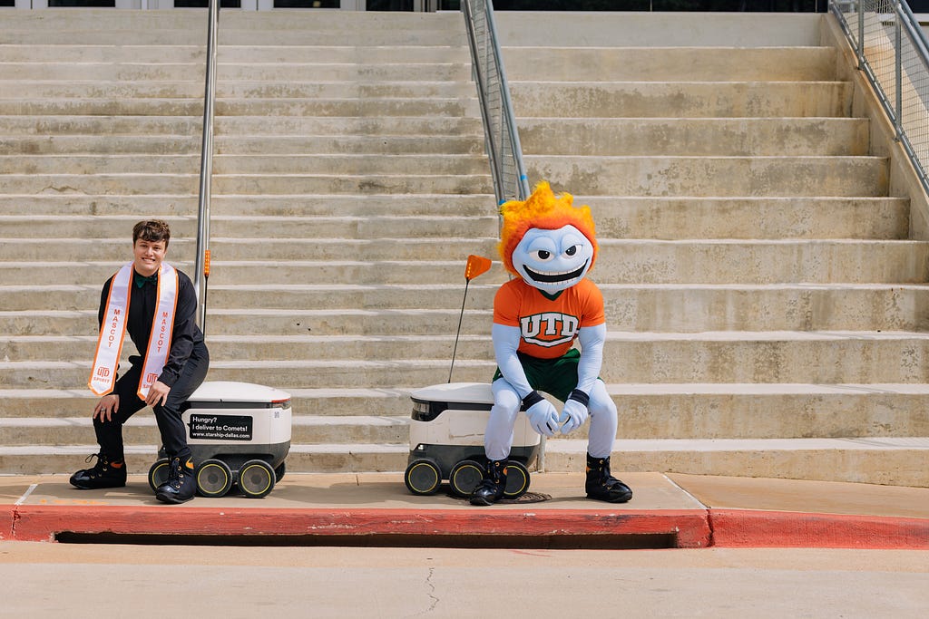 Eric and University of Texas Dallas Mascot, Temoc, posing with two of our Robots for graduation photos. Eric is dressed in black and Temoc is popping in oragne and green.