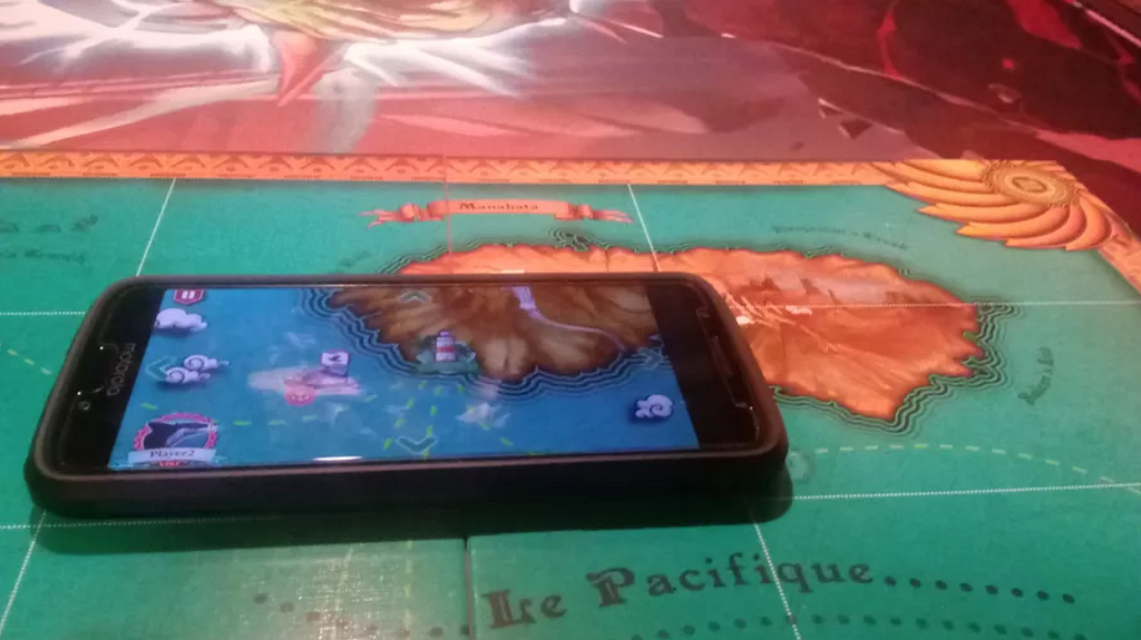A smartphone is placed on a game board depicting an island on an ocean labelled “Le Pacifique” that is divided into rectangular spaces. The phone screen appears to match what is beneath it on the board and shows some other icons, including a ship that represents the player. The phone is overlapping the space it is on, possibly due to its case.