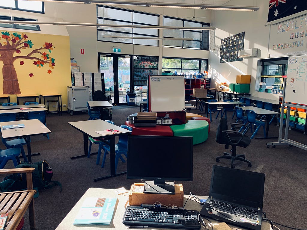 A desk with computer and laptop sits in the front of the picture. Behind, you can see a classroom with tables, chairs.