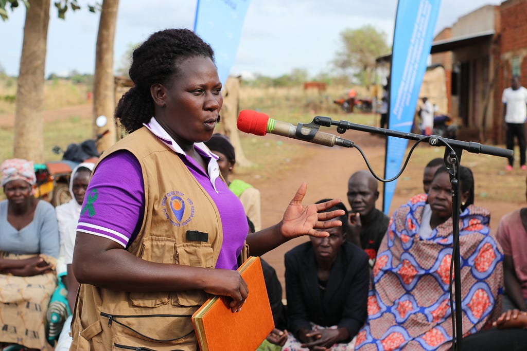 A black woman wearing a purple shirt and a brown jacket with the UN Trust Fund logo is talking through a microphone in front of a small crowd of people outside.